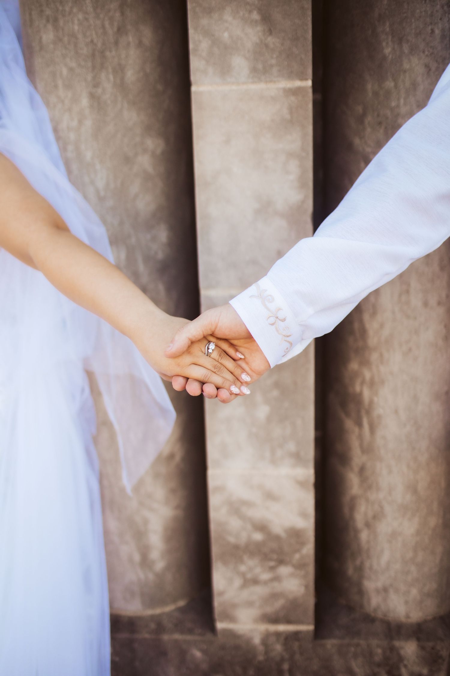 Close up of couple holding hands showing wedding rings against textured wall.