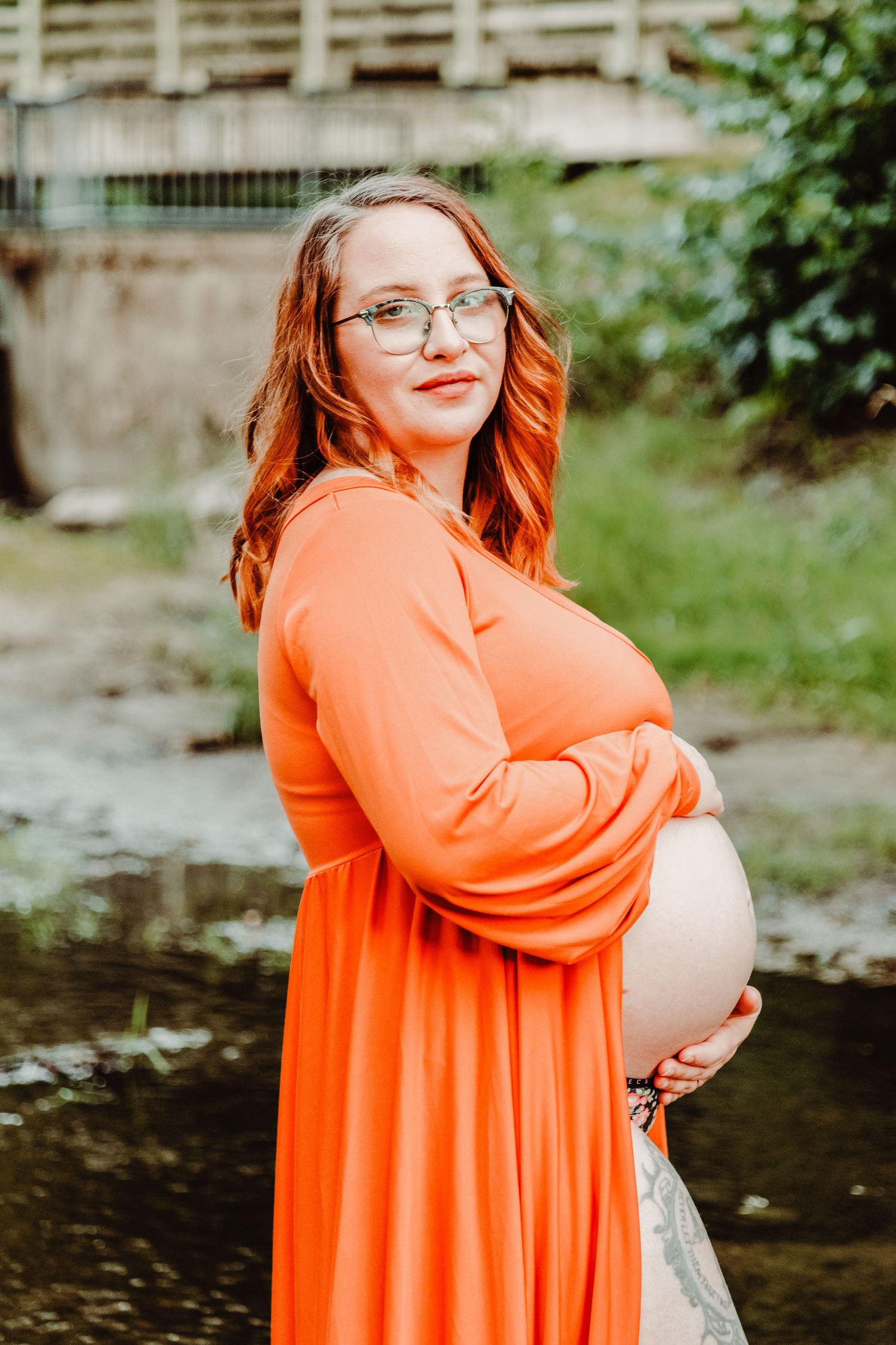 Joyful maternity portrait in bright coral dress by a peaceful stream.