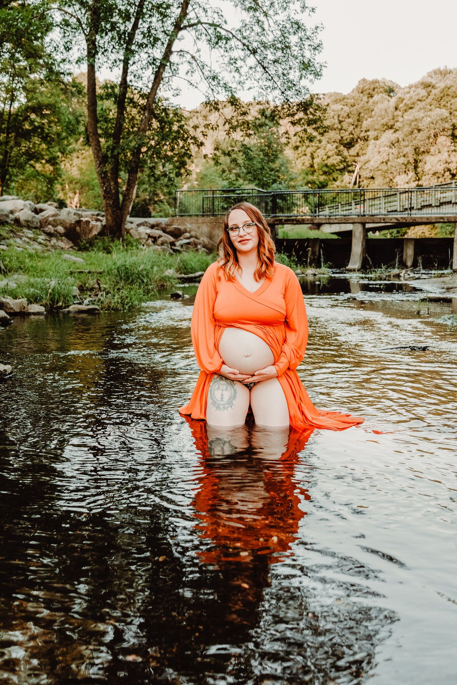Artistic maternity photo with orange dress reflection in stream water near rustic bridge.