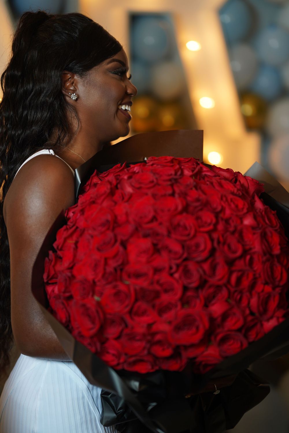 Someone wearing white holding an enormous bouquet of vibrant red roses against illuminated decor.