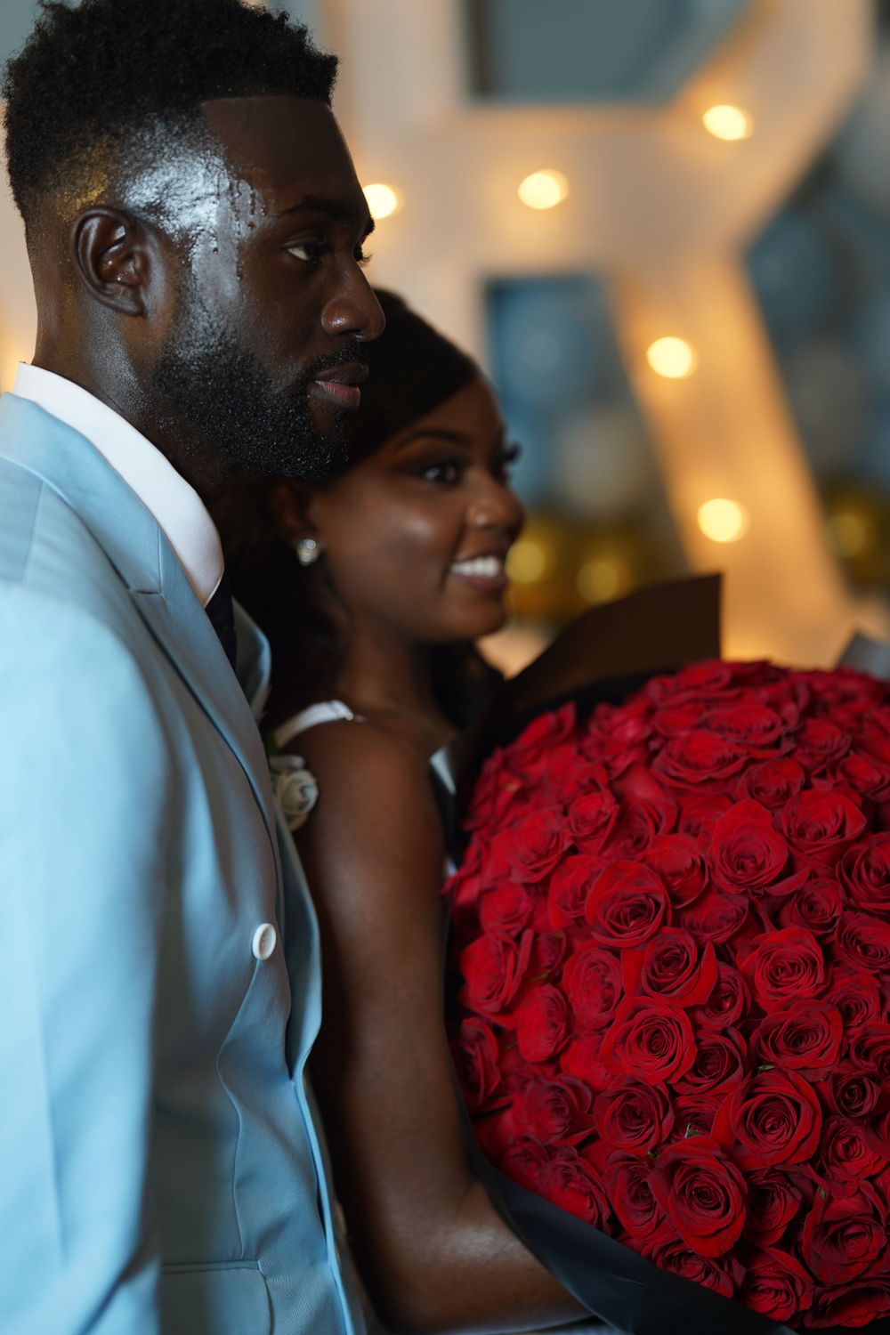 Couple posing with a stunning bouquet of red roses against an illuminated backdrop.