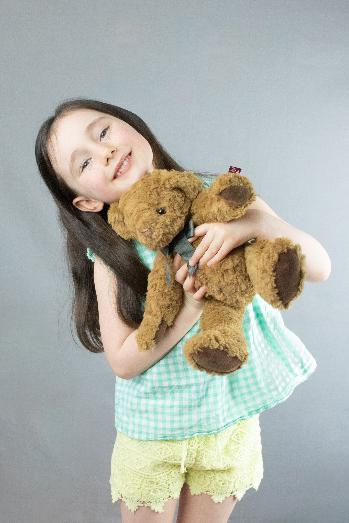 child photoshoot holding a teddy bear while smiling and tipping her head to the side wearing yellow shorts green top