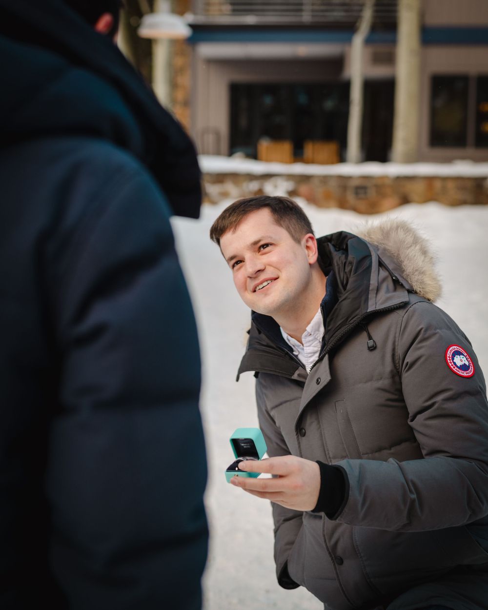 Person in winter coat smiles while holding small box outdoors in snowy setting.