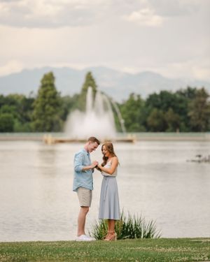 A couple celebrates their engagement by a lakeside fountain with mountains in the distance.