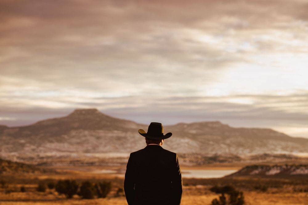 Man in black suit and cowboy hat stands alone gazing at a vast golden desert landscape.