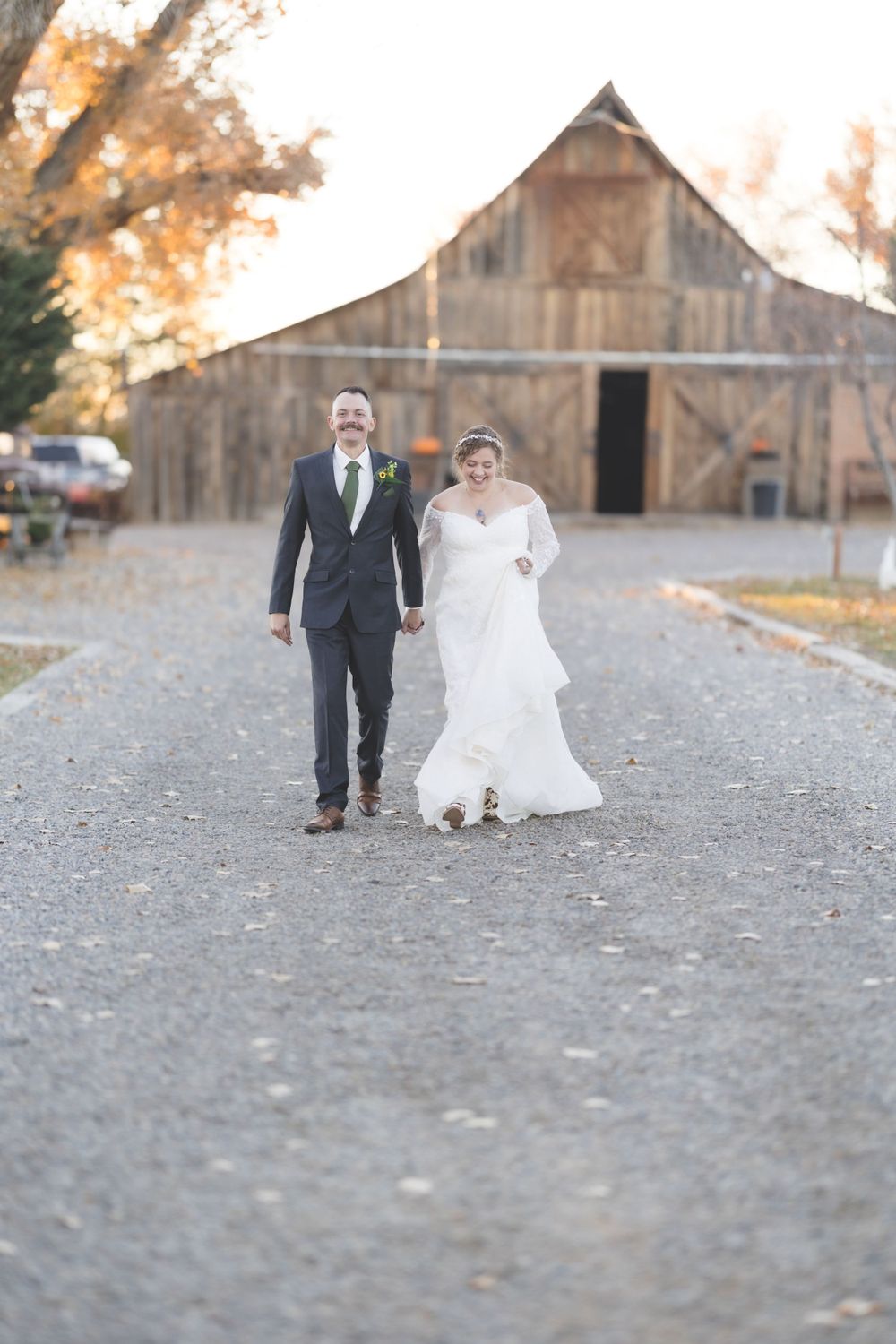 Smiling bride and groom walk hand in hand down a gravel path with rustic barn backdrop.