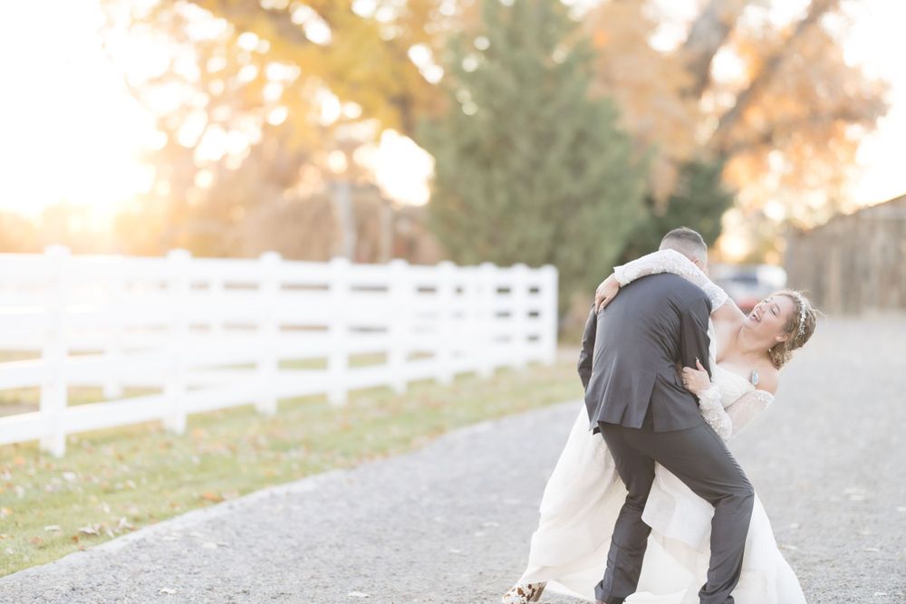 Groom dips bride for a kiss along a tree-lined road with white fence at golden hour.