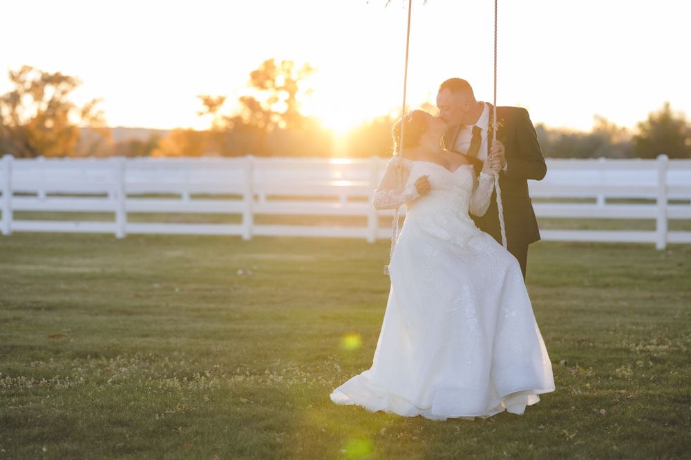 Bride and groom share a kiss on a rope swing at sunset beside a white fence on a farm.