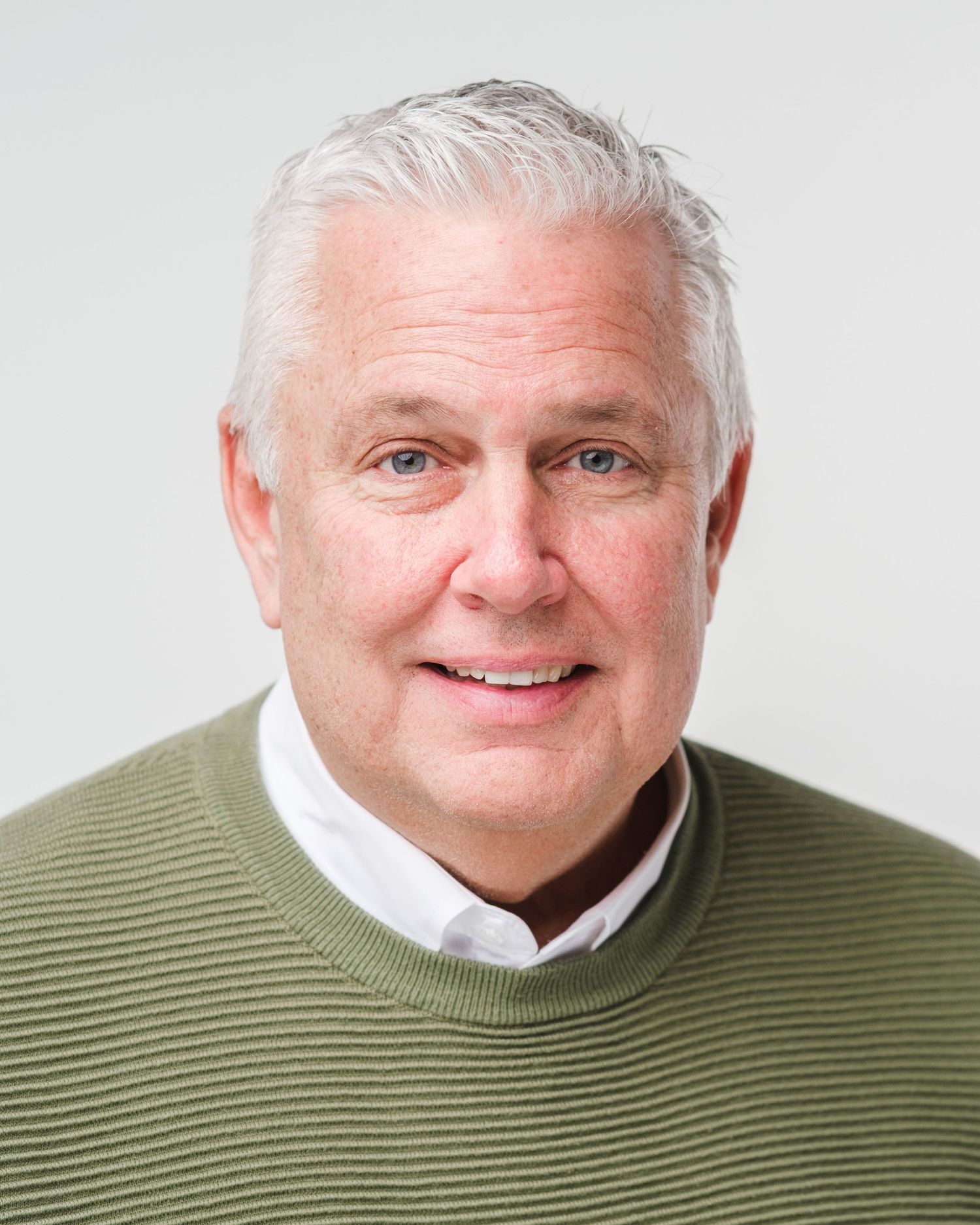 Professional headshot of a person wearing an olive green sweater over a white collared shirt against a light background.