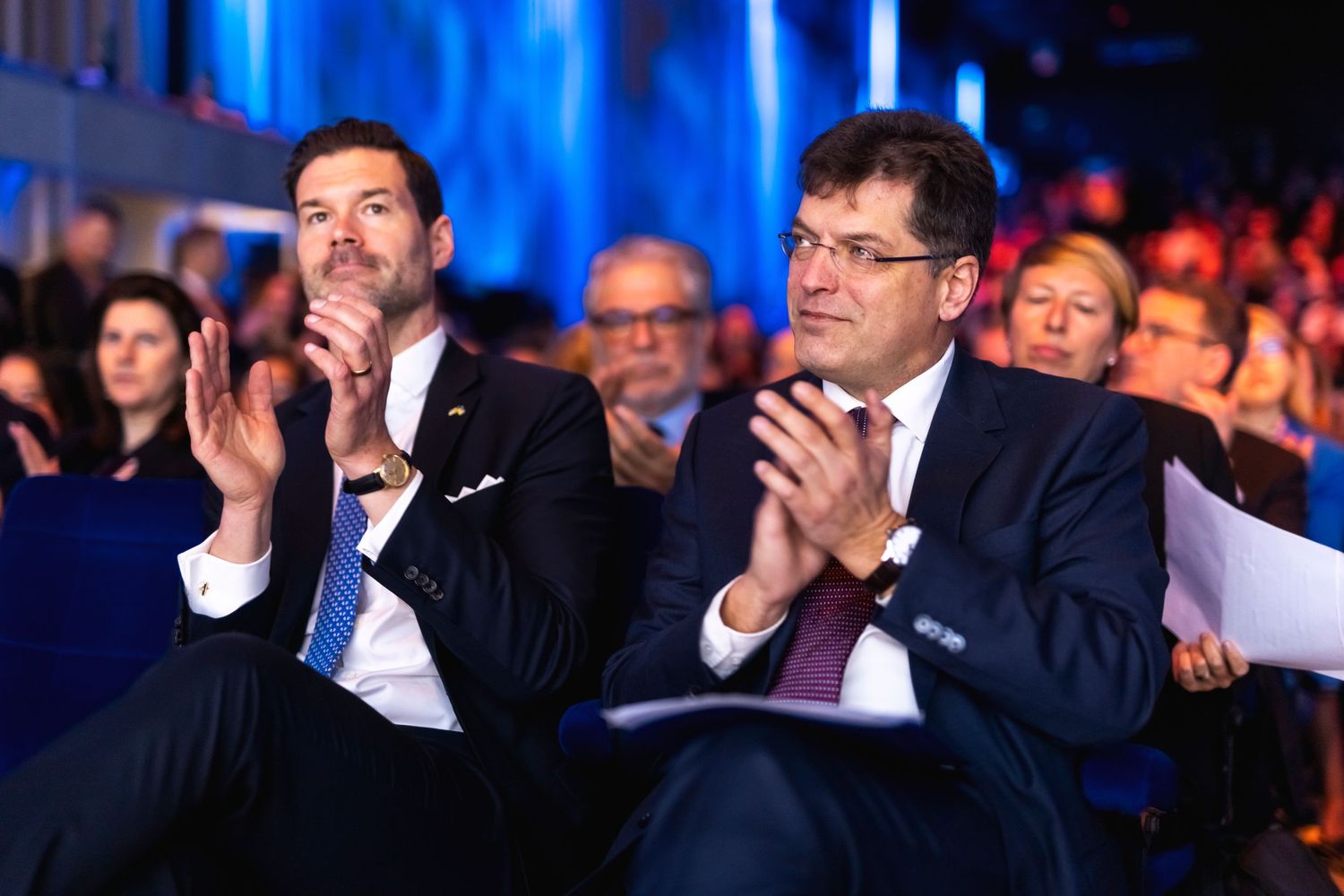 Conference attendees clapping during presentation in blue-lit auditorium.