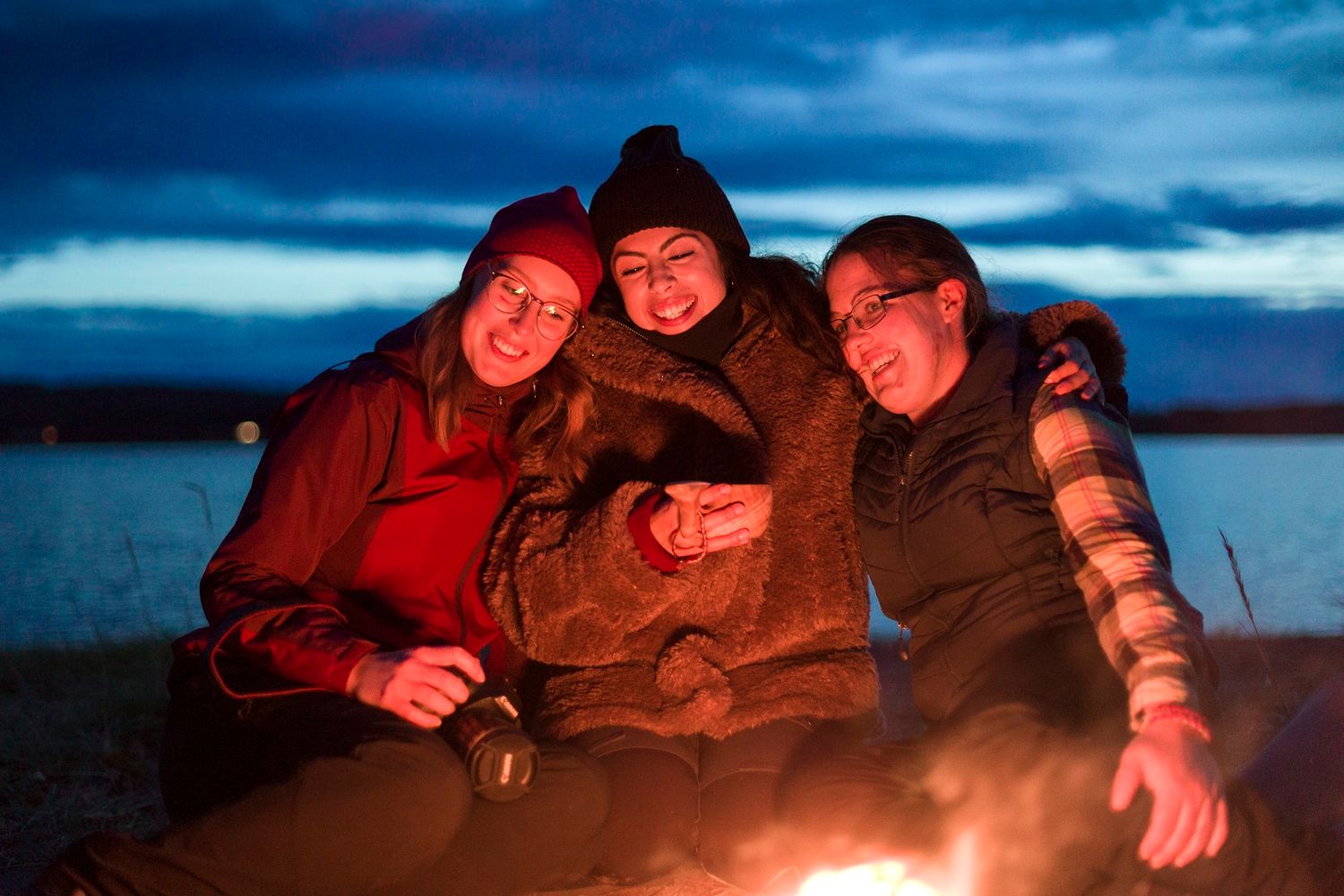 Three friends smiling together by a warm campfire near a lake at dusk.