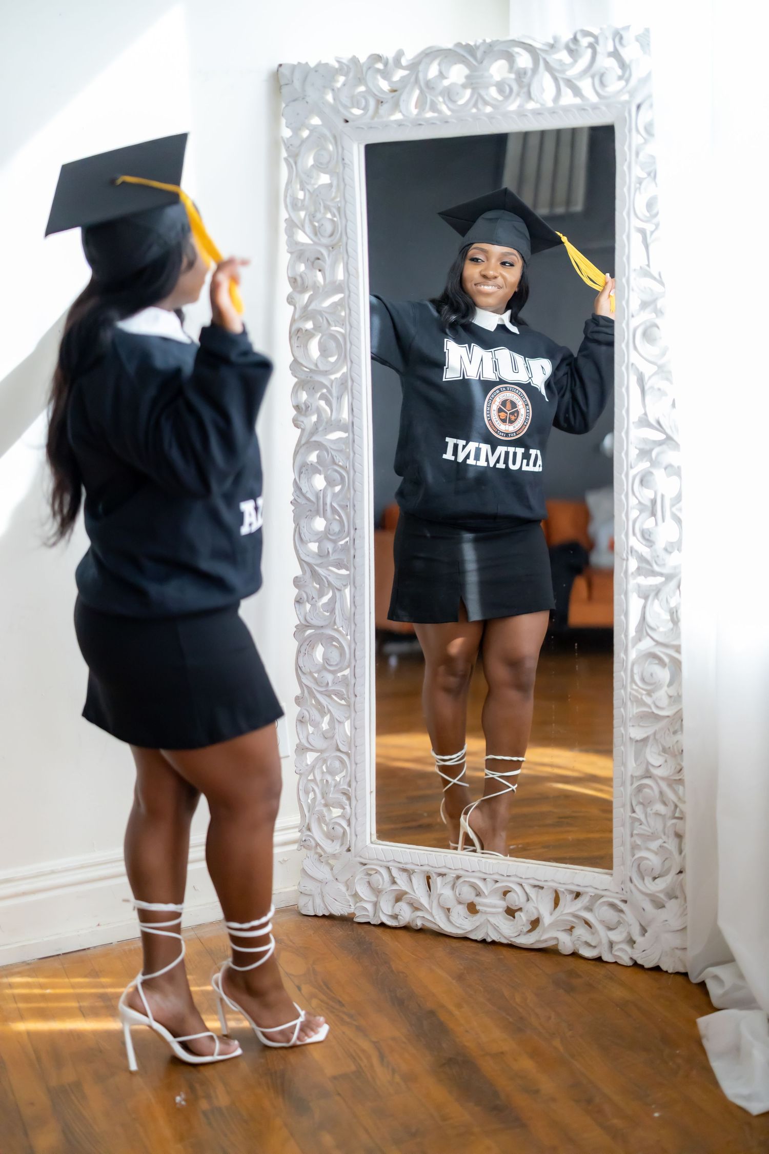 Graduate taking mirror selfie in ornate white frame mirror wearing cap and gown.