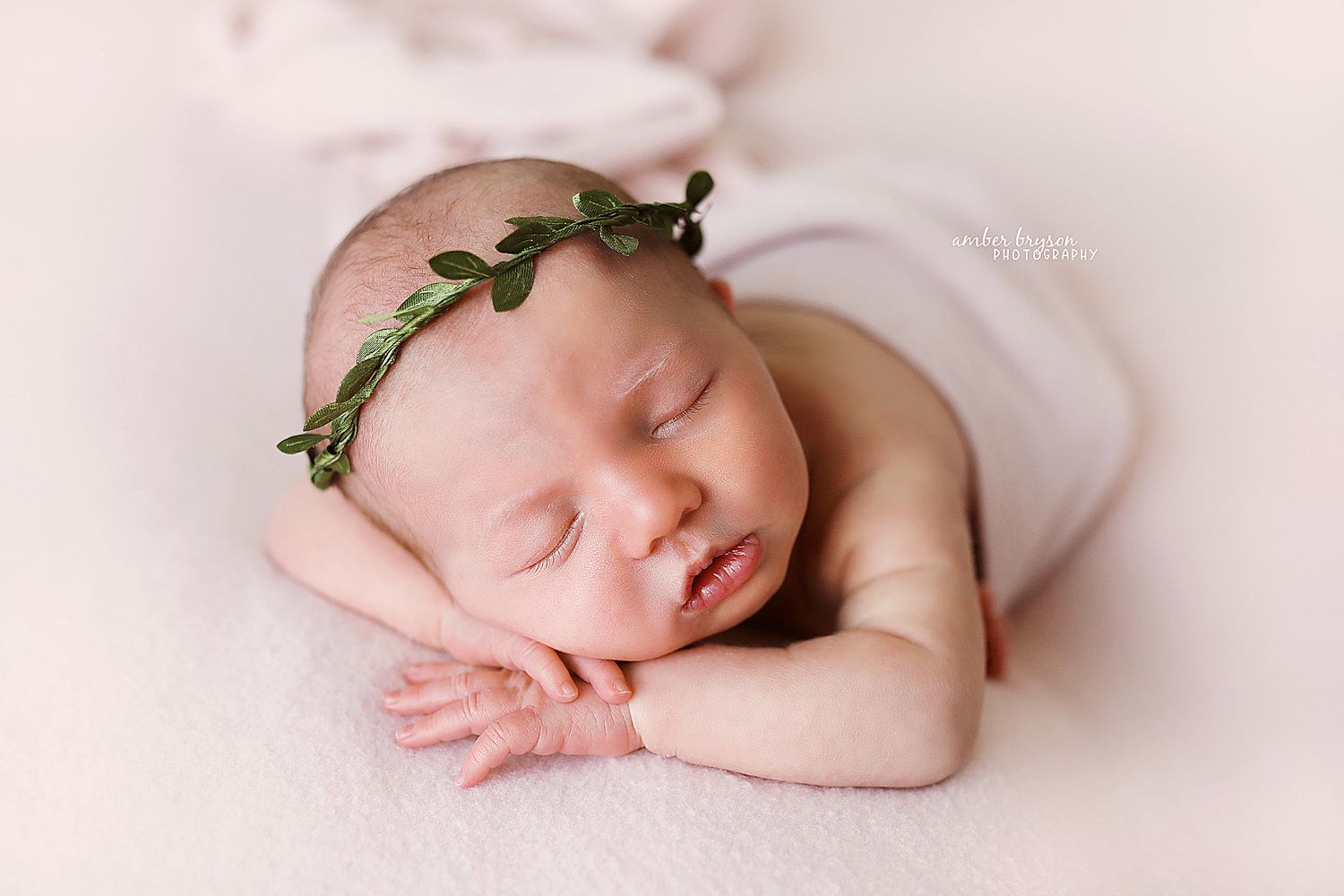 Sleeping newborn wearing delicate leaf crown on soft pink background.