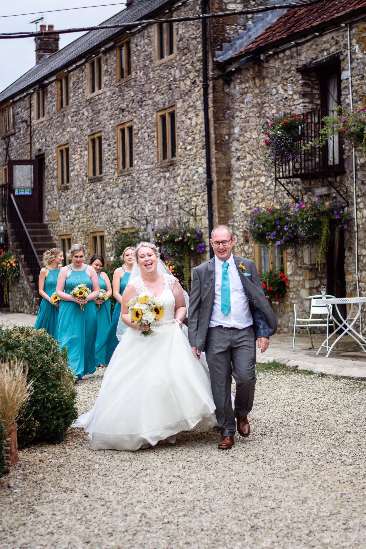 Wedding party walks along path by rustic stone buildings with colorful flowers.