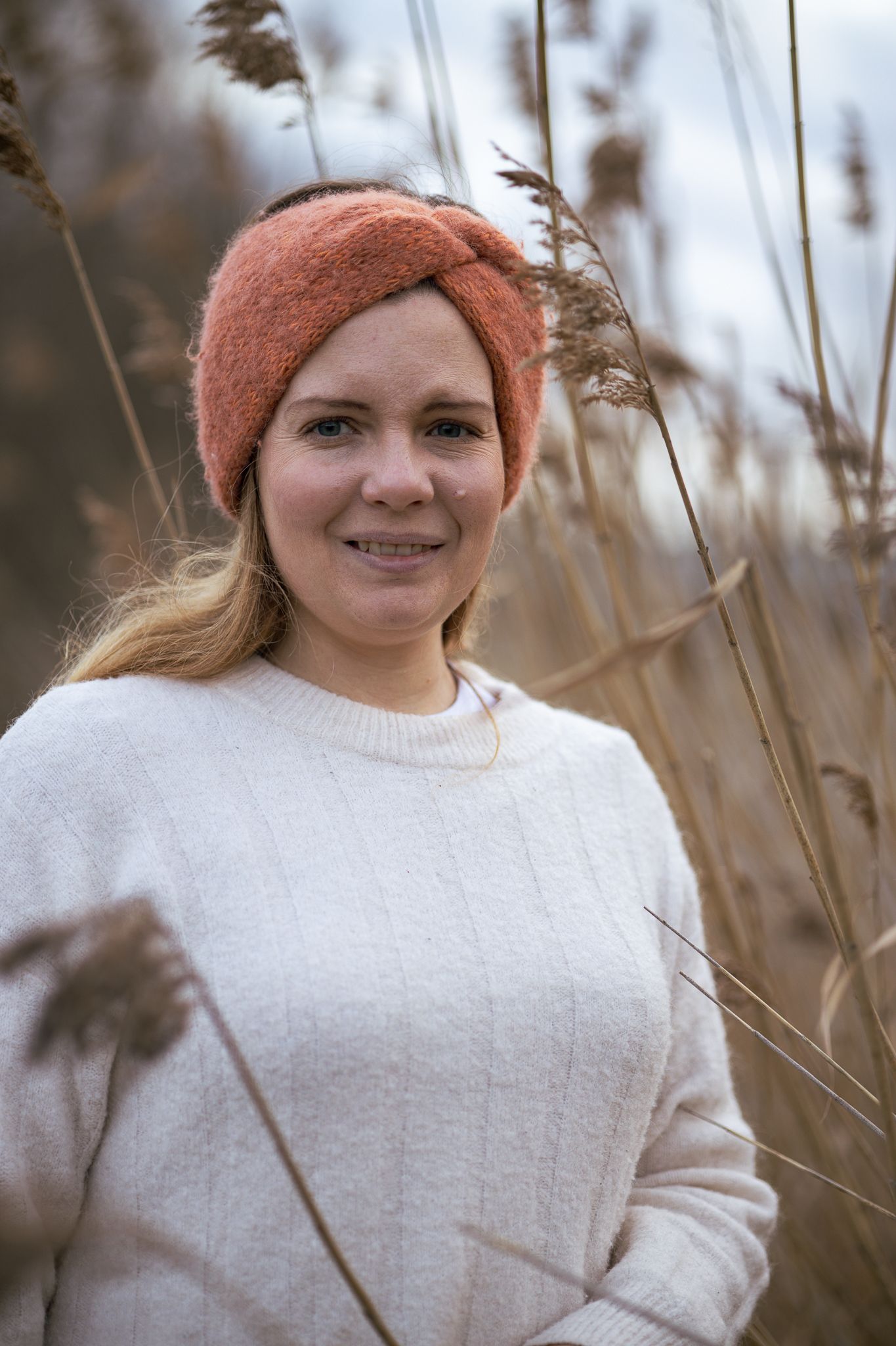 A smiling person wearing a coral knit headband and cream sweater poses among tall grass in an autumn field.