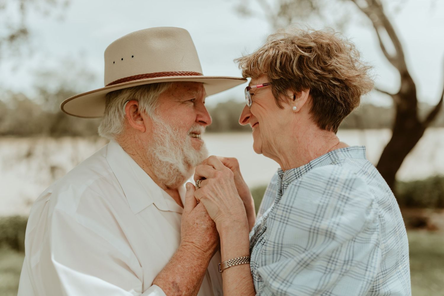 An elderly couple shares a tender moment outdoors under a wide-brimmed hat.