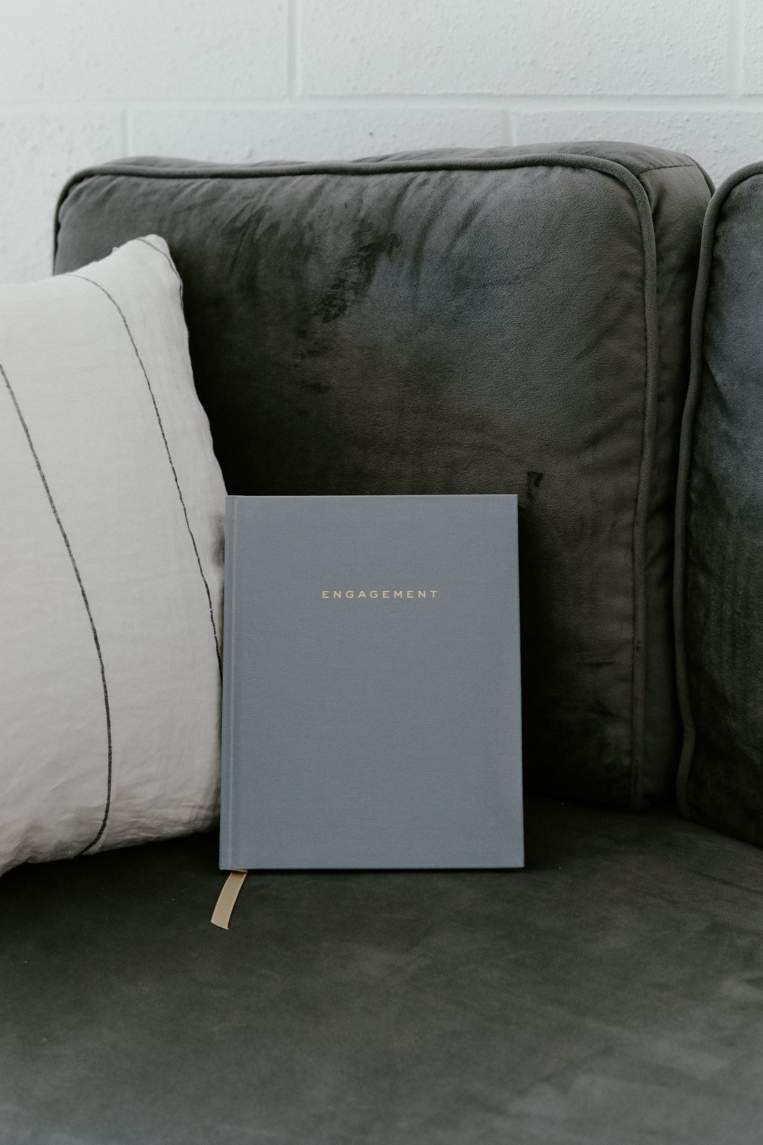A gray engagement journal sits on a dark cushioned couch with white pillows.