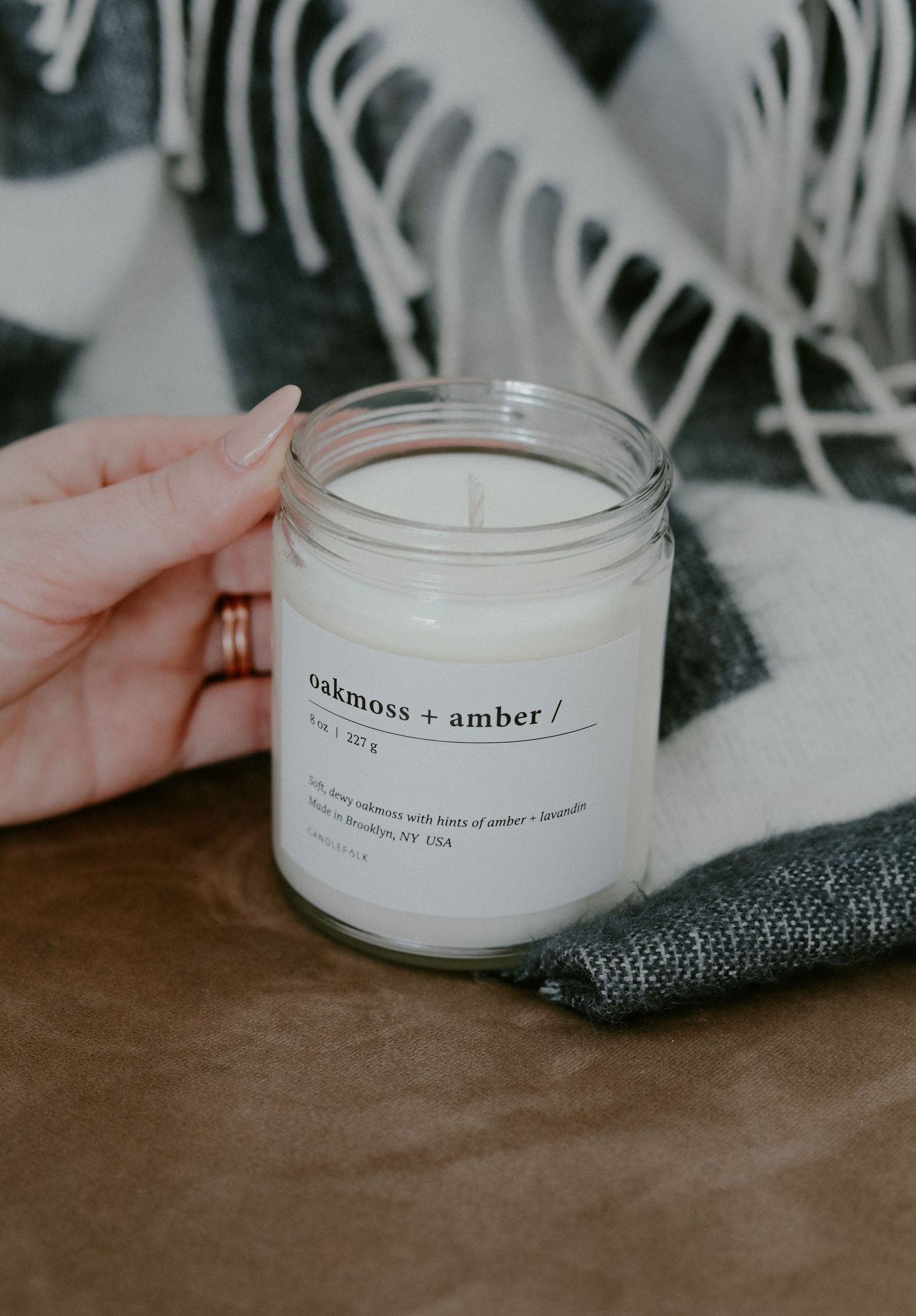 Hand holding a white scented candle jar labeled oakmoss and amber against patterned background.
