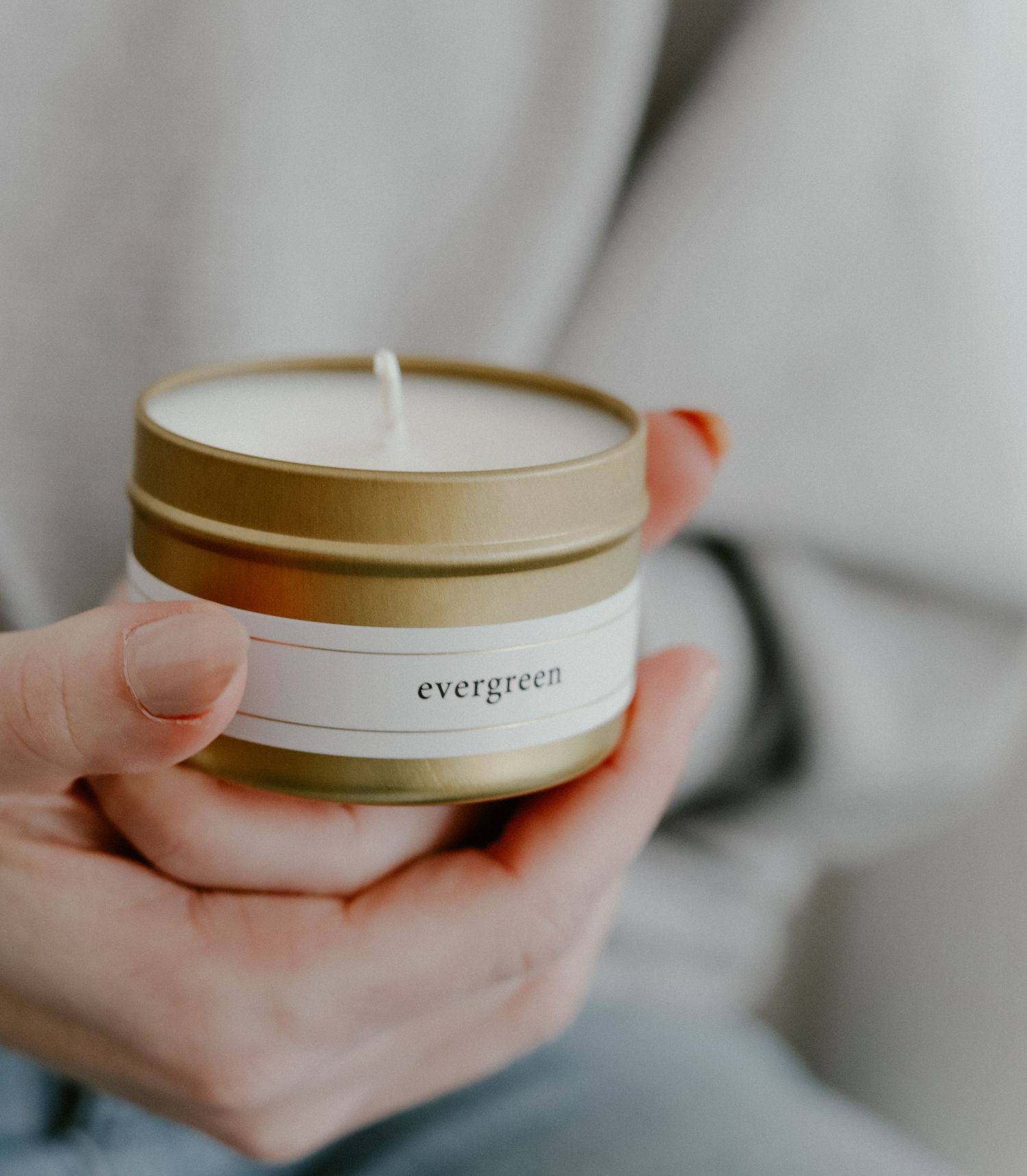 Gold tin candle labeled 'evergreen' held in hand against light background.