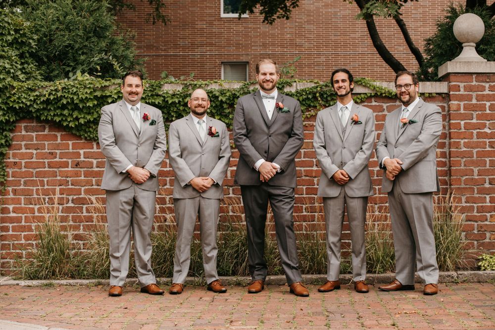 Five groomsmen in matching gray suits with boutonnieres pose in a line against a brick wall with greenery.