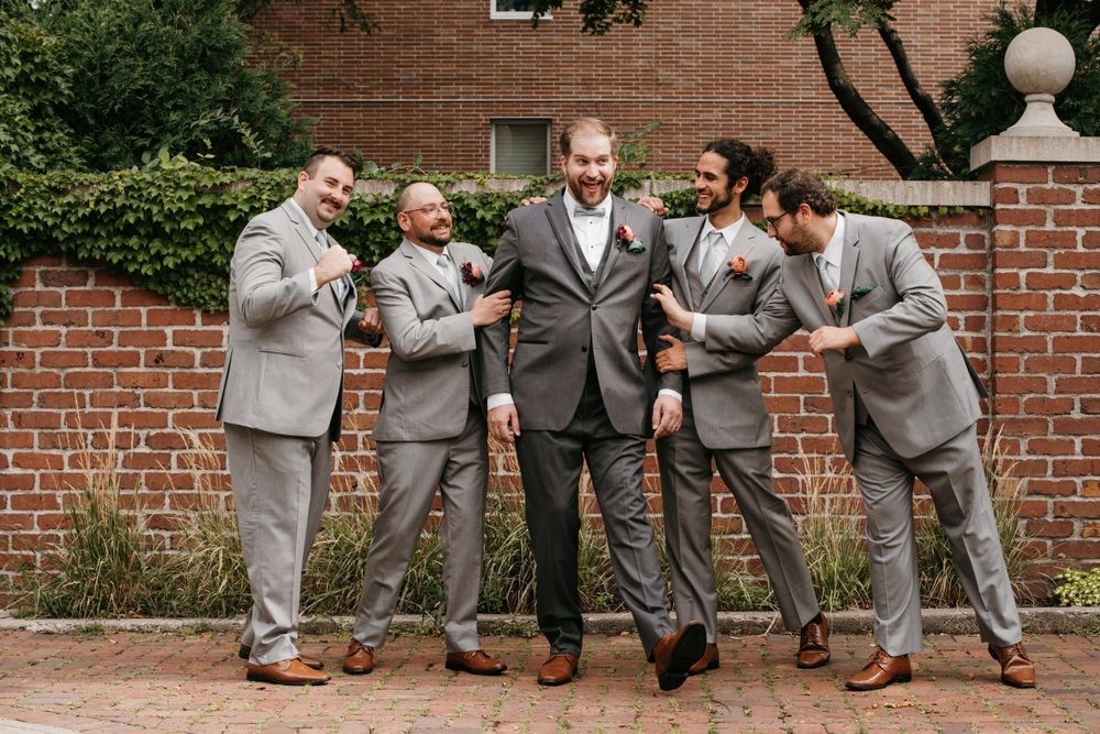Groom and groomsmen laughing together in gray suits with boutonnieres against a brick wall.