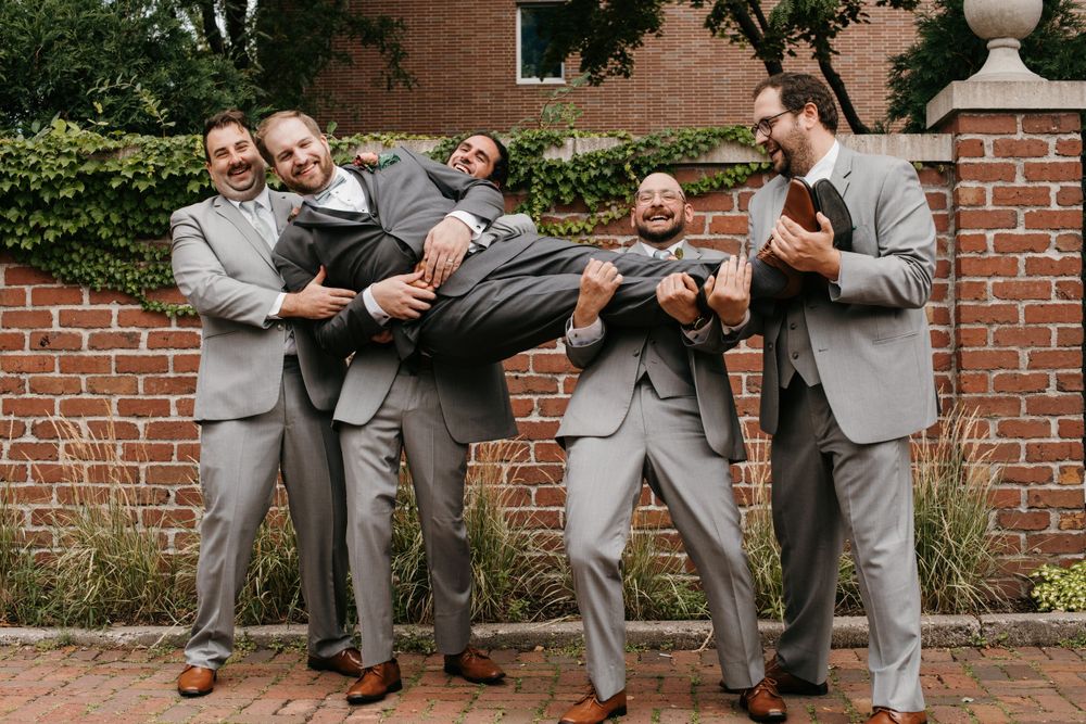 Groomsmen playfully lifting the groom in gray suits outside a brick wall at a wedding.