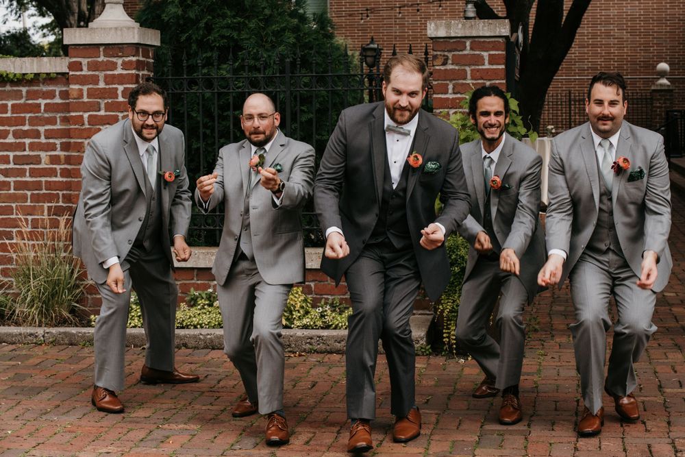 Groom and groomsmen dancing and celebrating in gray suits on a brick patio.