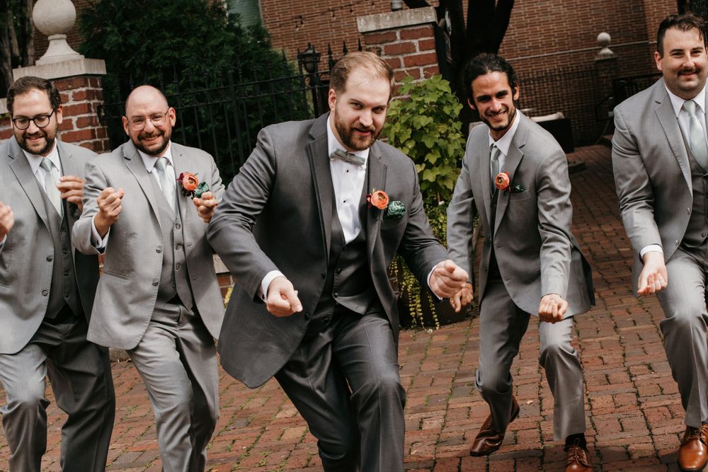 Groom and groomsmen joyfully dancing together in gray suits at a wedding celebration.