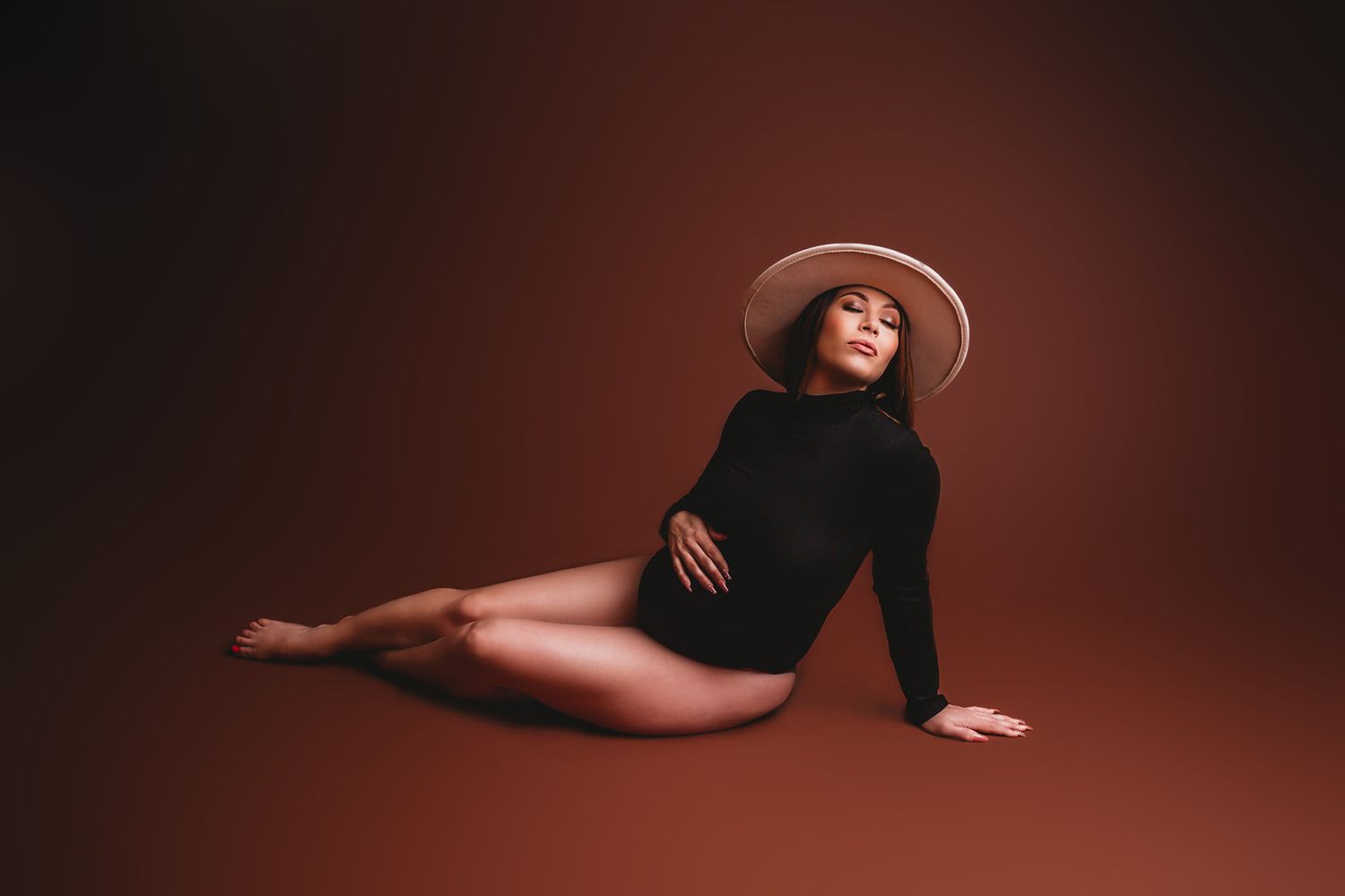 Studio maternity photo featuring a pose in black leotard and wide brim hat against dark backdrop.