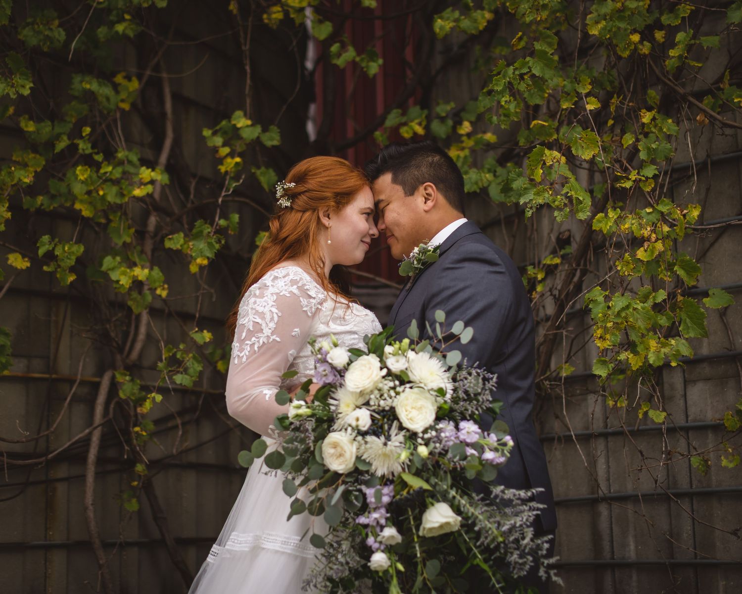 Wedding couple shares intimate moment with bride holding elegant white floral bouquet against ivy wall.