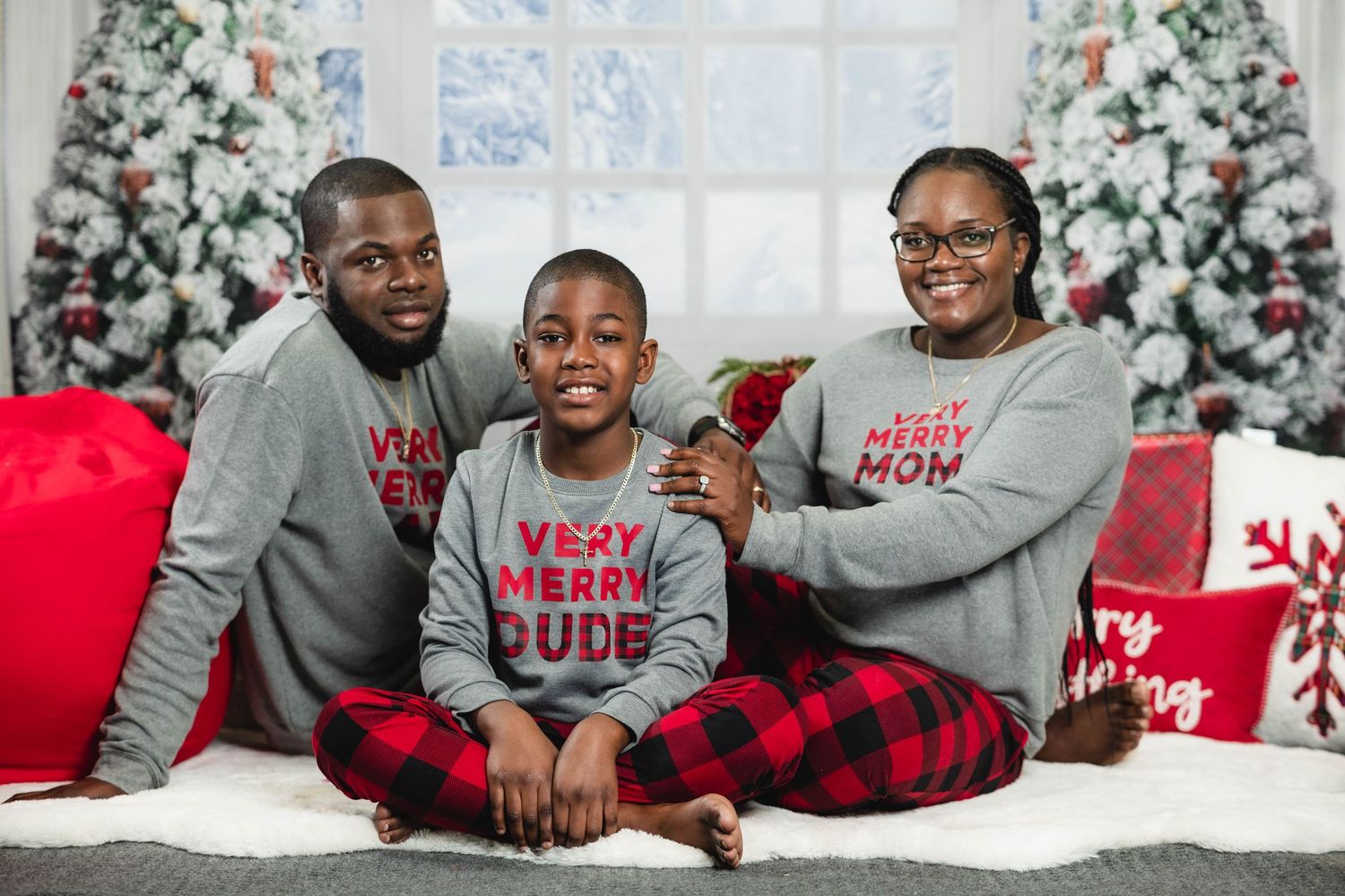 Family of three in matching gray and red plaid Christmas pajamas posing by holiday tree.