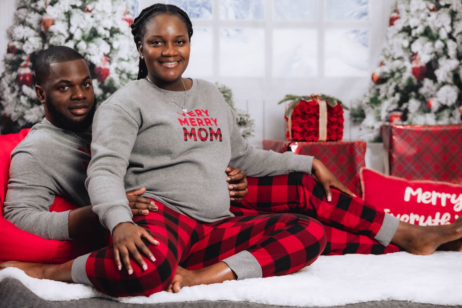 Pregnant couple in matching Christmas pajamas posing in front of decorated holiday tree with gifts.