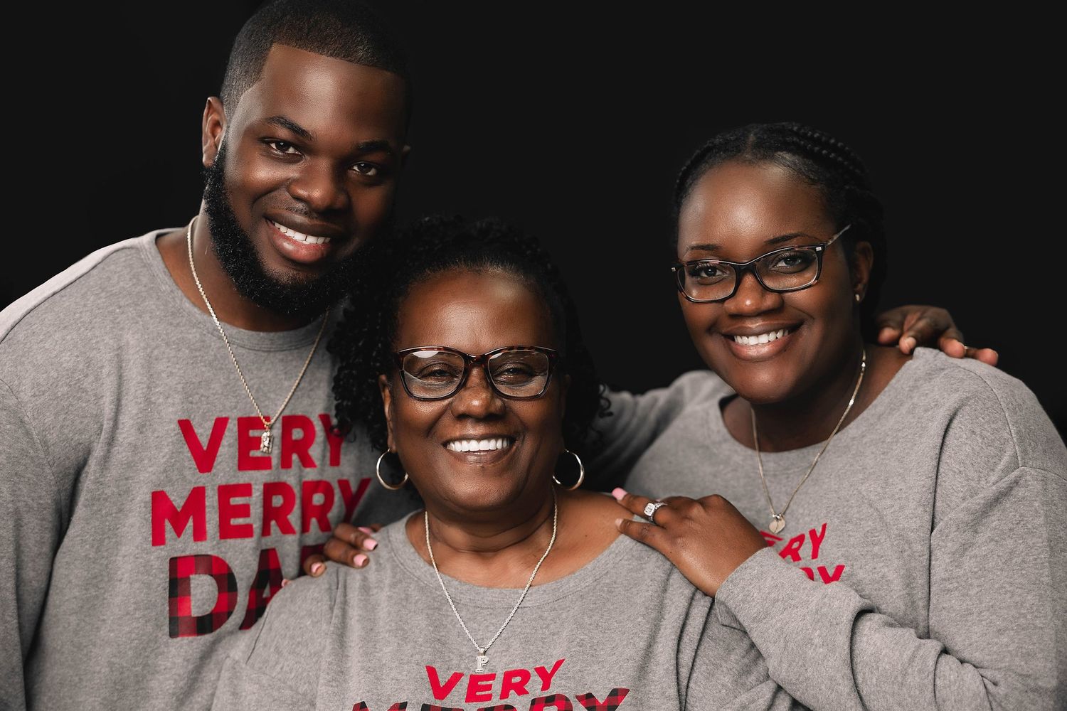 Three people smiling in matching grey Very Merry Days holiday sweatshirts against dark background.