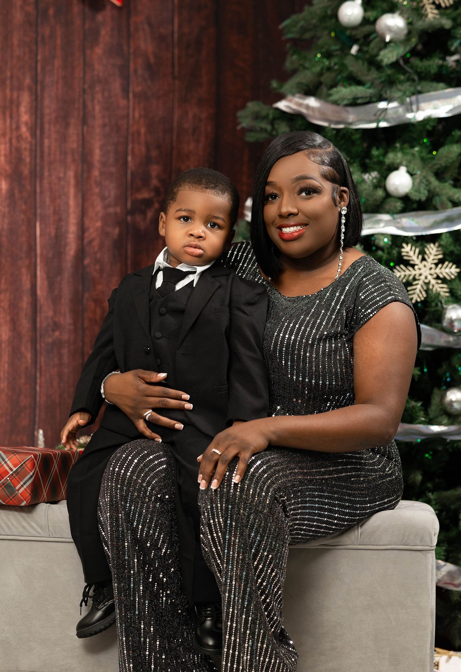 Mother and toddler son in matching black formal outfits posing beside Christmas tree.
