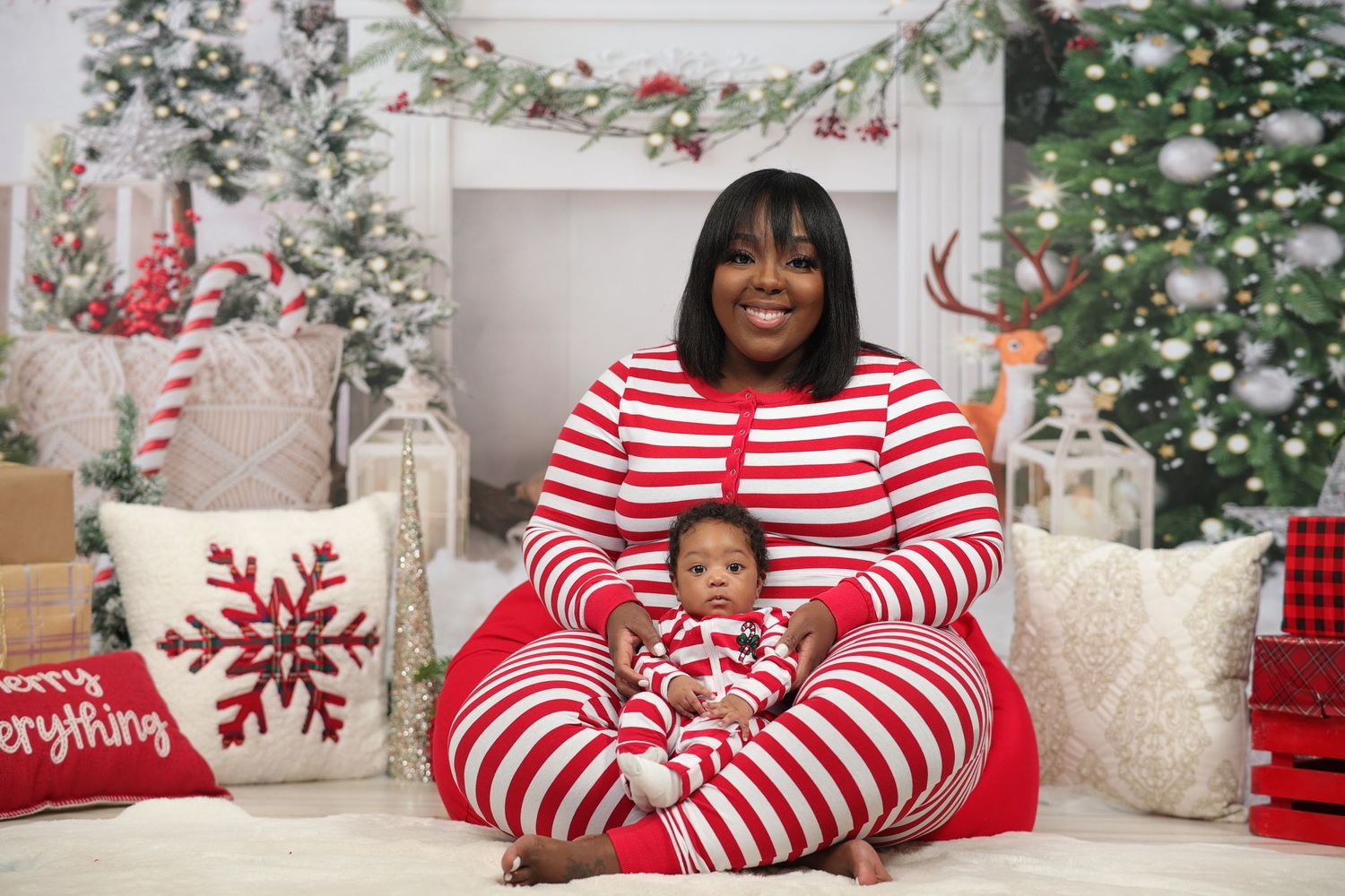 Mother in red striped pajamas sitting with newborn baby in matching outfit by Christmas decor.