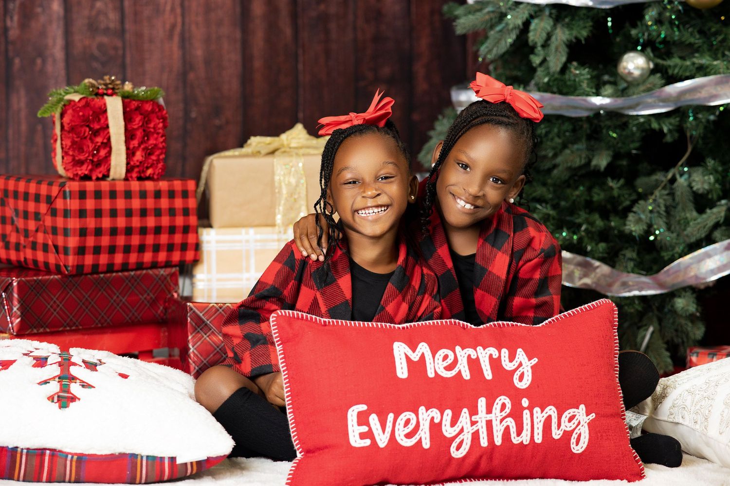 Two girls in red plaid outfits hugging by a Christmas tree.