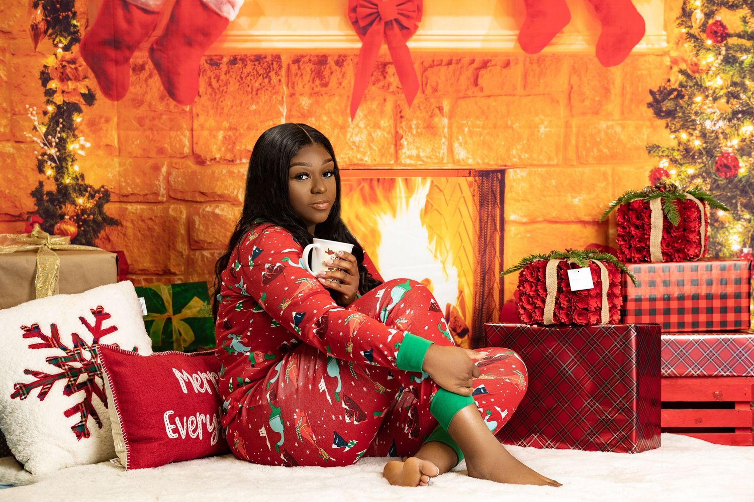 Young woman in Christmas pajamas sitting by fireplace holding mug with holiday gifts and decorations.