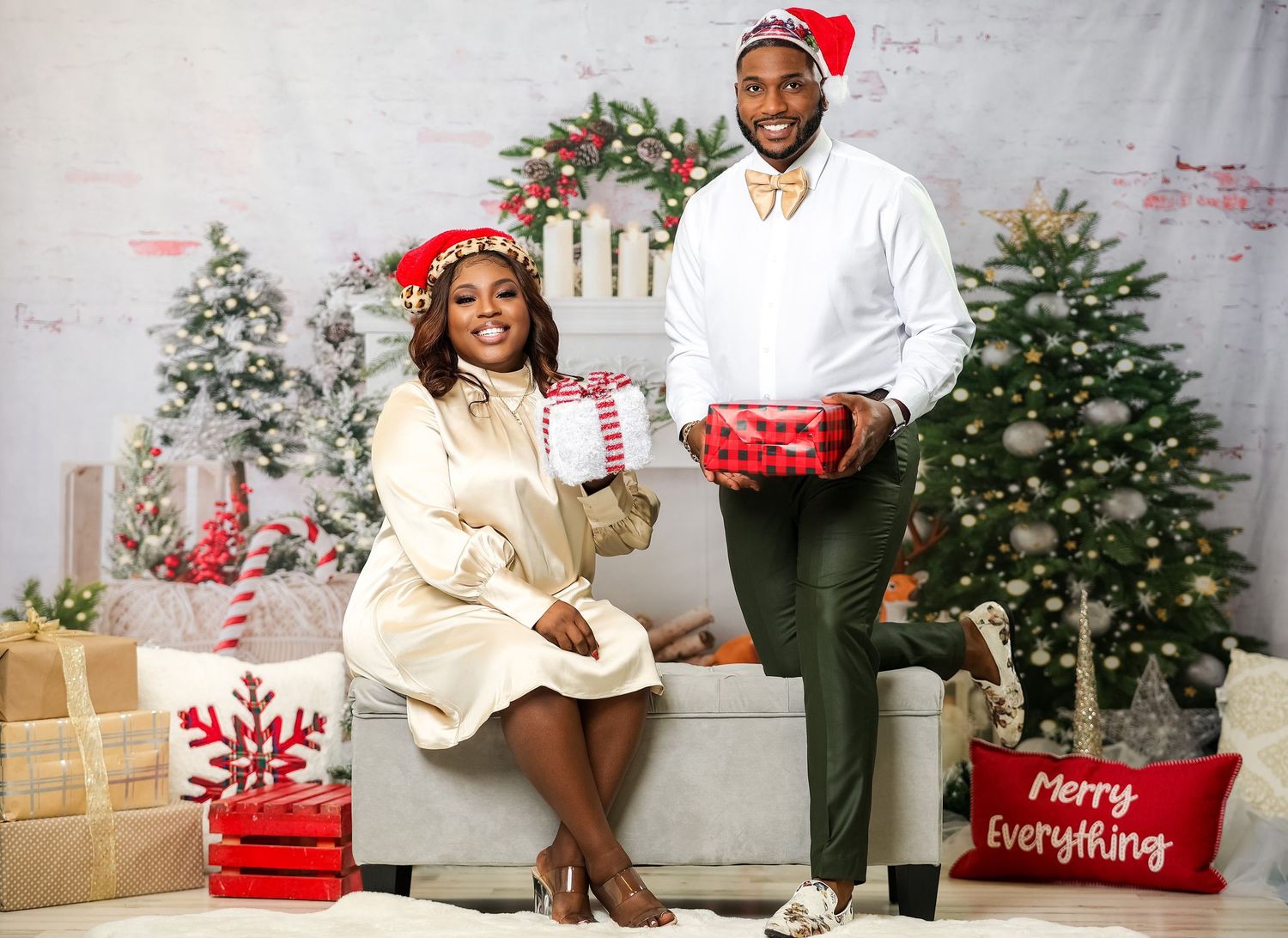 Couple in Christmas attire posing with gifts near decorated tree, wearing Santa hats in festive setting.
