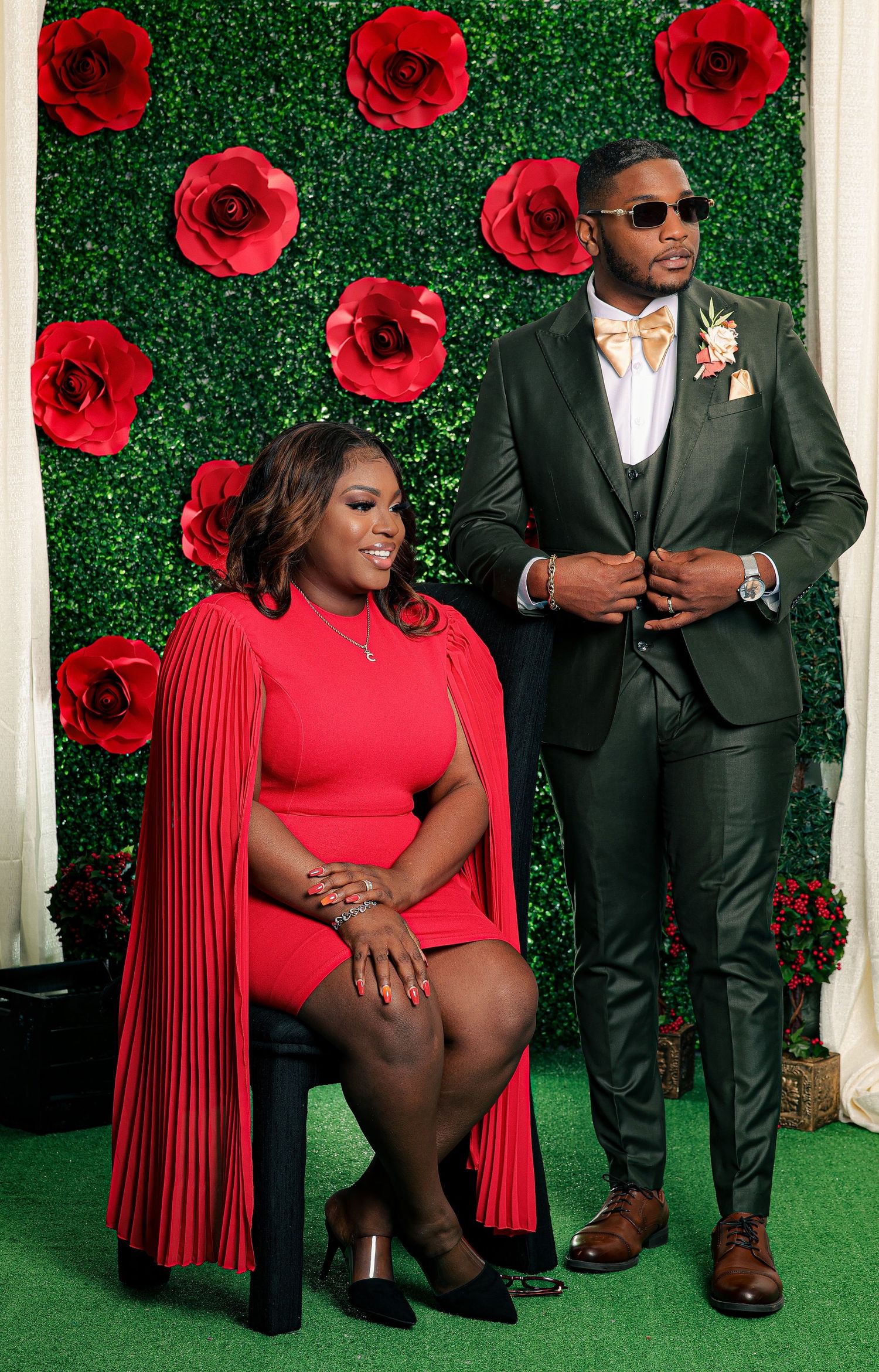 Elegant couple posing in front of red roses backdrop, woman in red cape dress seated, man in dark green suit standing.