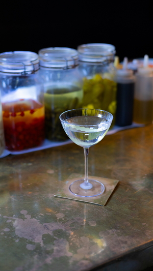 A sophisticated martini cocktail on a rustic table with various mixers and garnishes in mason jars behind it.