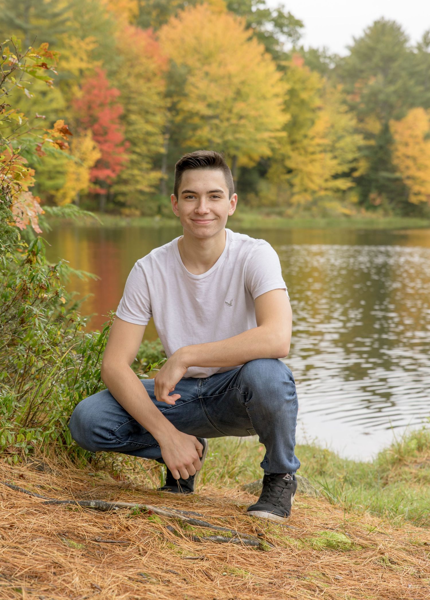 senior photography fall color young man white tee and jeans squatting down in front of a pond Rochester NH