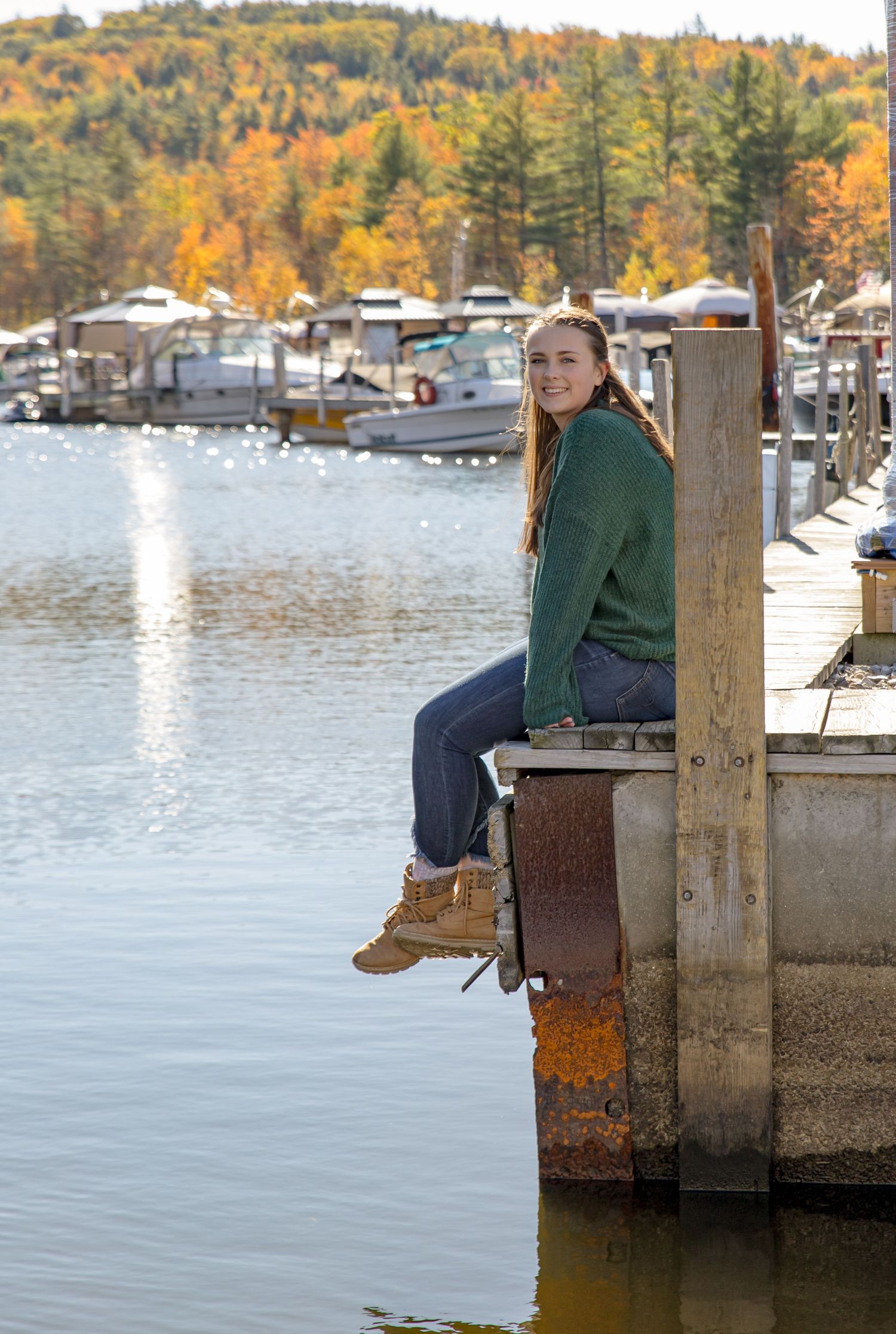 young high school senior sitting on the town docks in Wolfeboro NH with fall foliage and lake Winnipesaukee