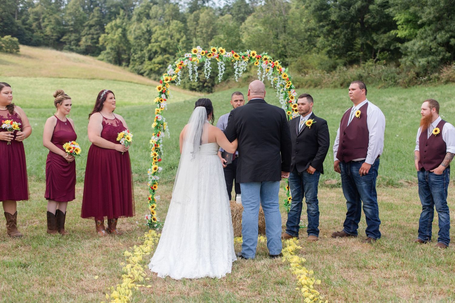 Wedding party lined up in burgundy attire with sunflower bouquets in a scenic field setting.