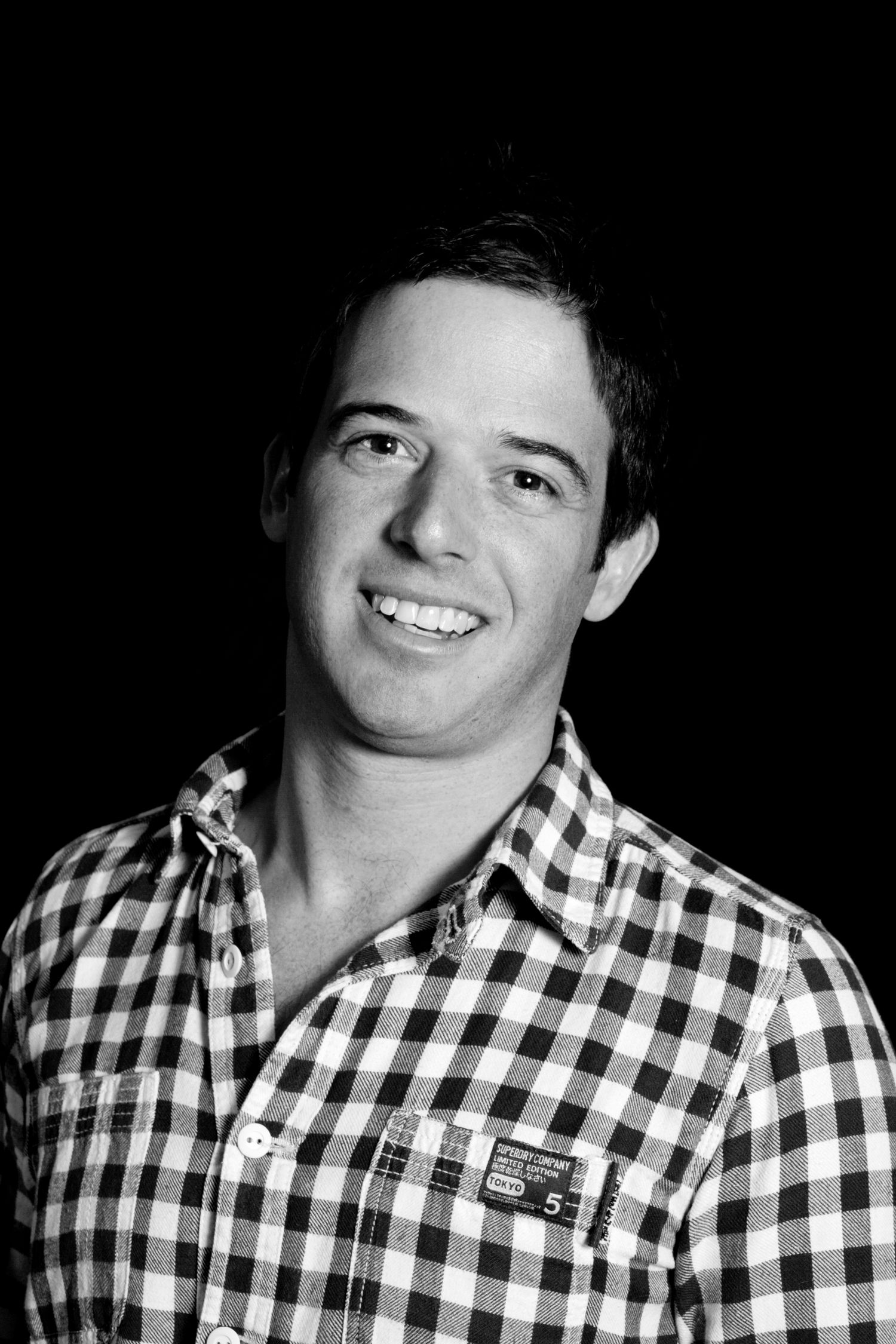 Black and white headshot of person wearing checkered shirt against dark background during business branding photoshoot.