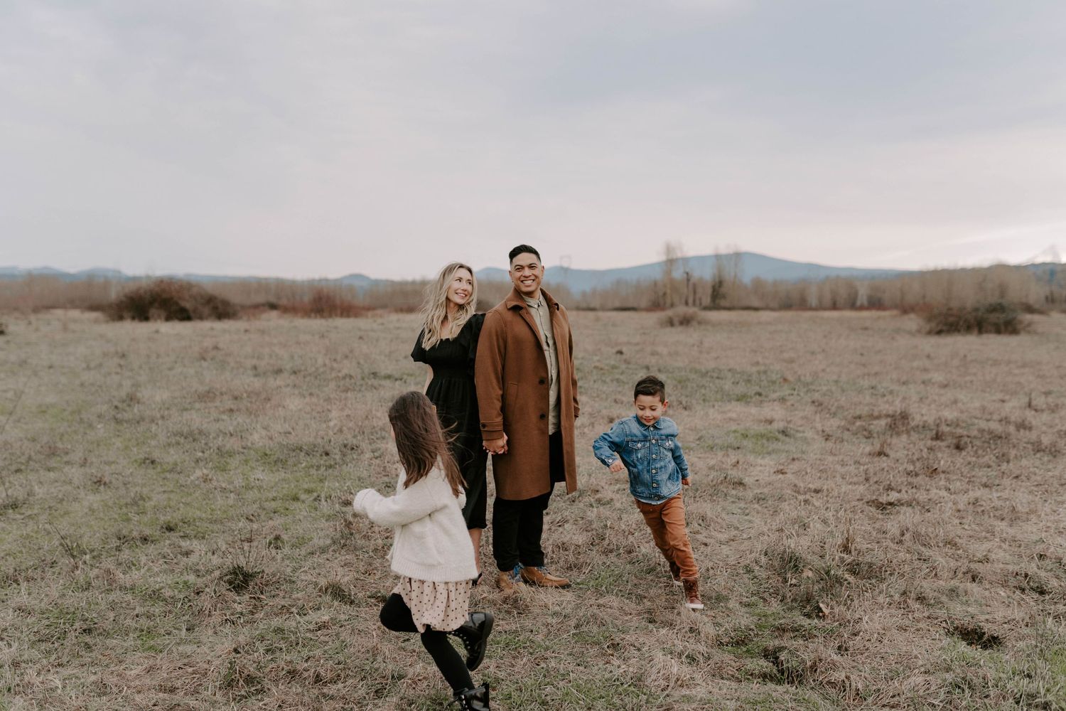 A family enjoys a playful moment together in a field with mountains visible in the distant background.