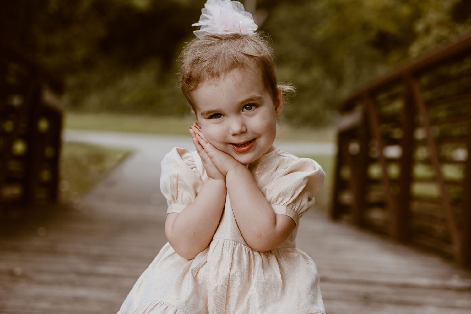 A child in a light dress strikes a playful pose with hands on their cheeks on a wooden bridge.
