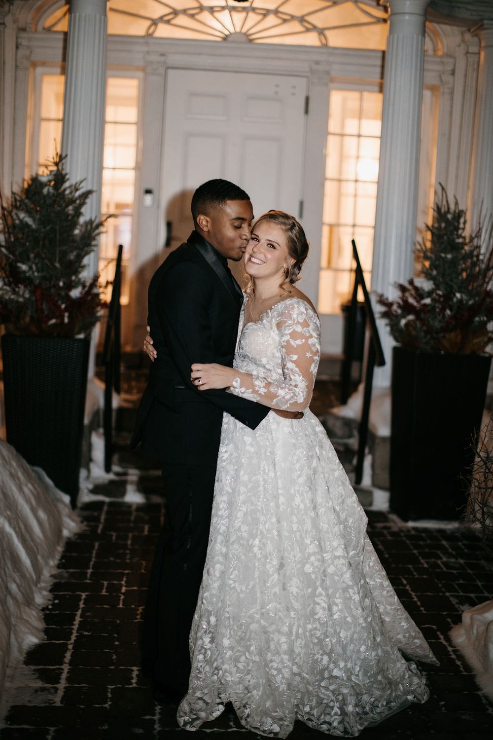 Bride and groom embrace at elegant wedding venue entrance with white columns and winter decor.