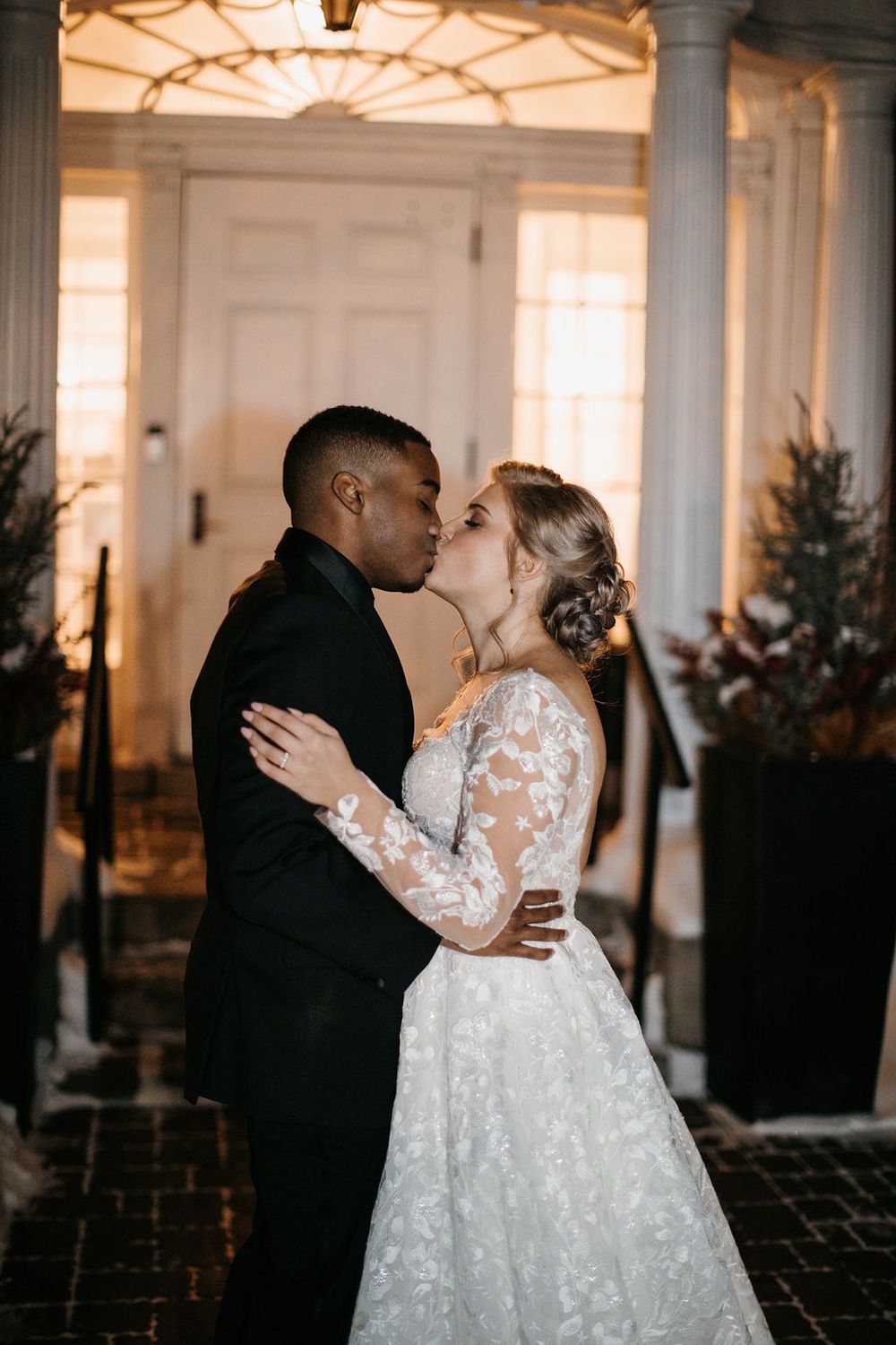 Bride and groom share tender kiss near white columned entrance of wedding venue at night.