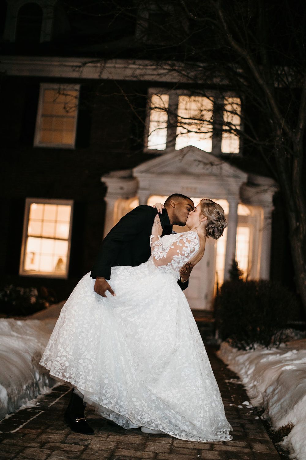 Groom dramatically dips bride for a kiss outside snow-covered wedding venue at night.