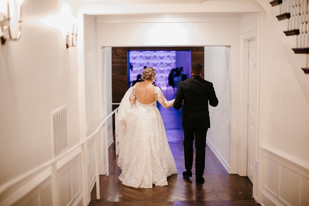 Bride and groom walking hand in hand through elegant white hallway toward glowing reception entrance.